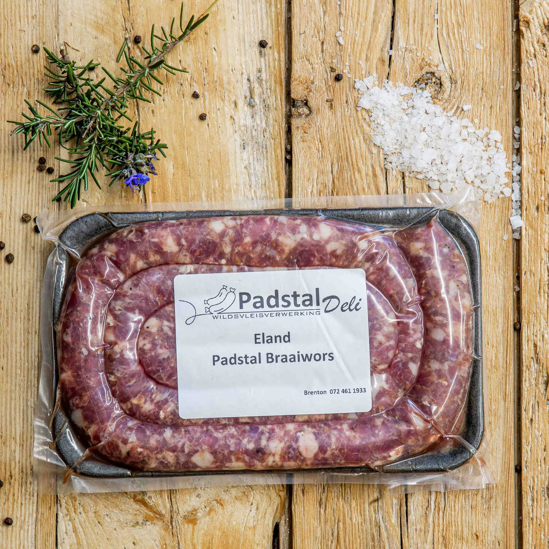 Padstal Deli Grabouw Braaiwors coiled in a tight spiral on a light wooden background, garnished with coarse sea salt, black peppercorns, and fresh rosemary.