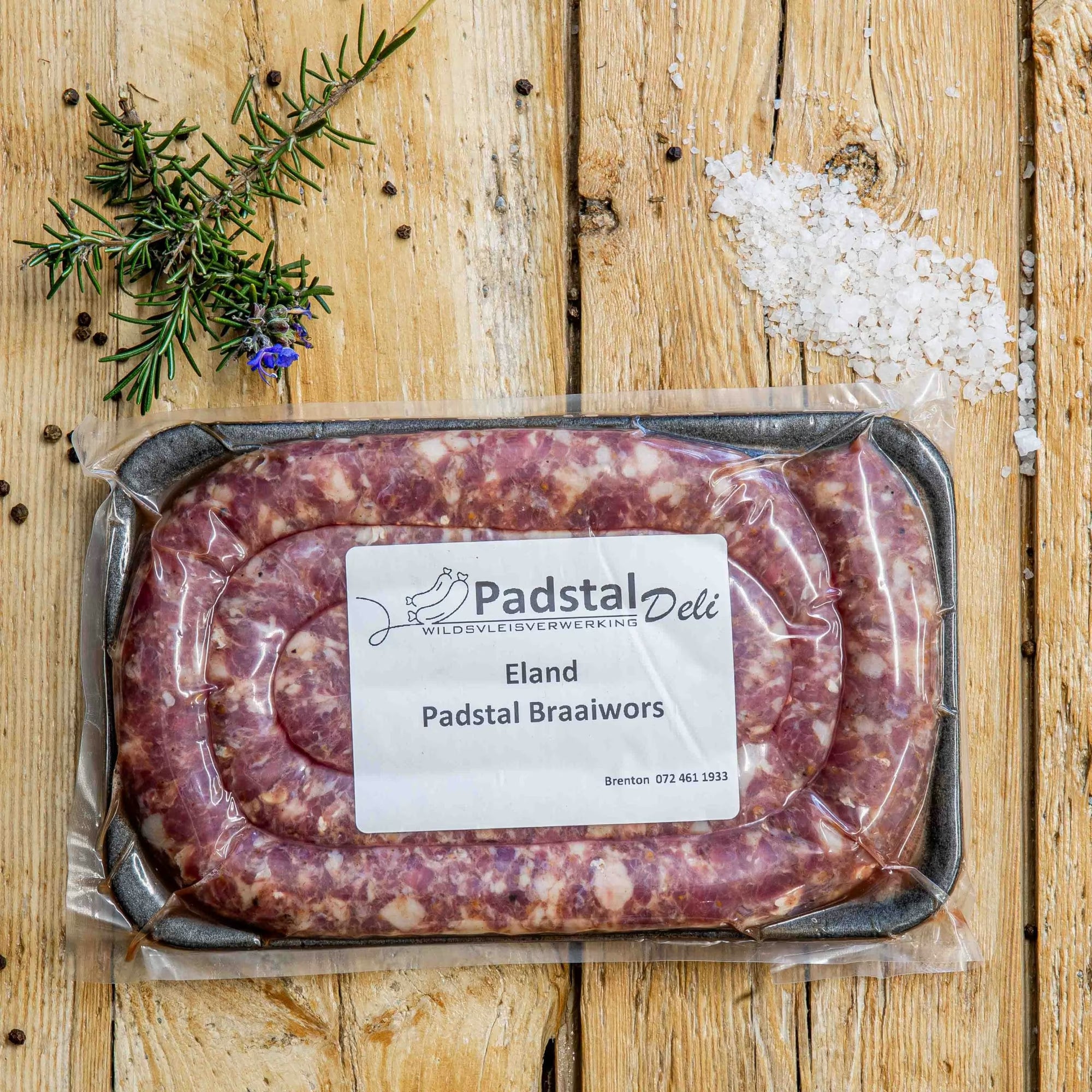 Padstal Deli vacuum-sealed Eland Padstal Braaiwors packaged on a black tray, displayed on a wooden background with coarse sea salt, peppercorns, and fresh rosemary.