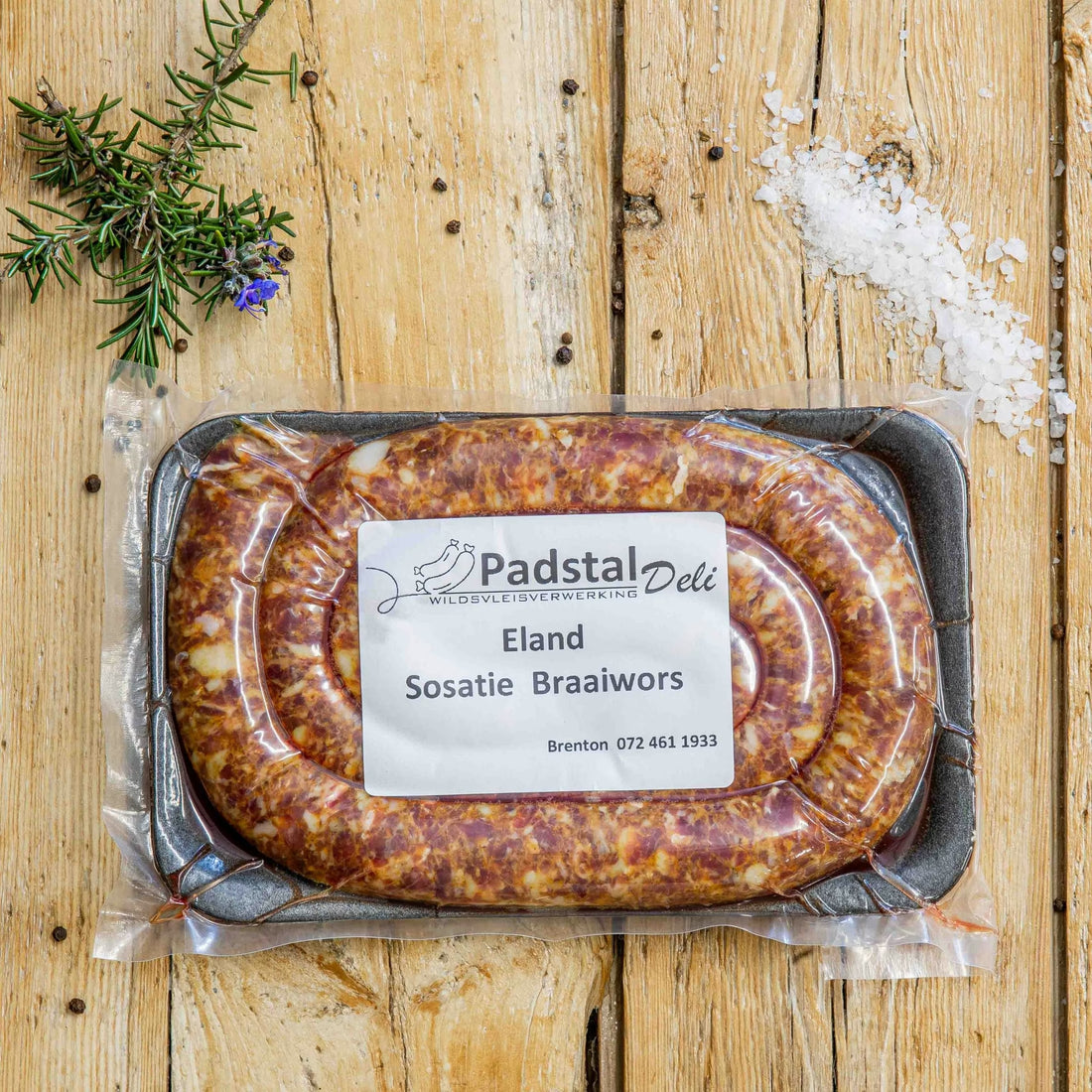 Padstal Deli Eland Sosatie Braaiwors coiled in a tight spiral on a light rustic wooden background, decorated with coarse sea salt, black peppercorns, and fresh rosemary.