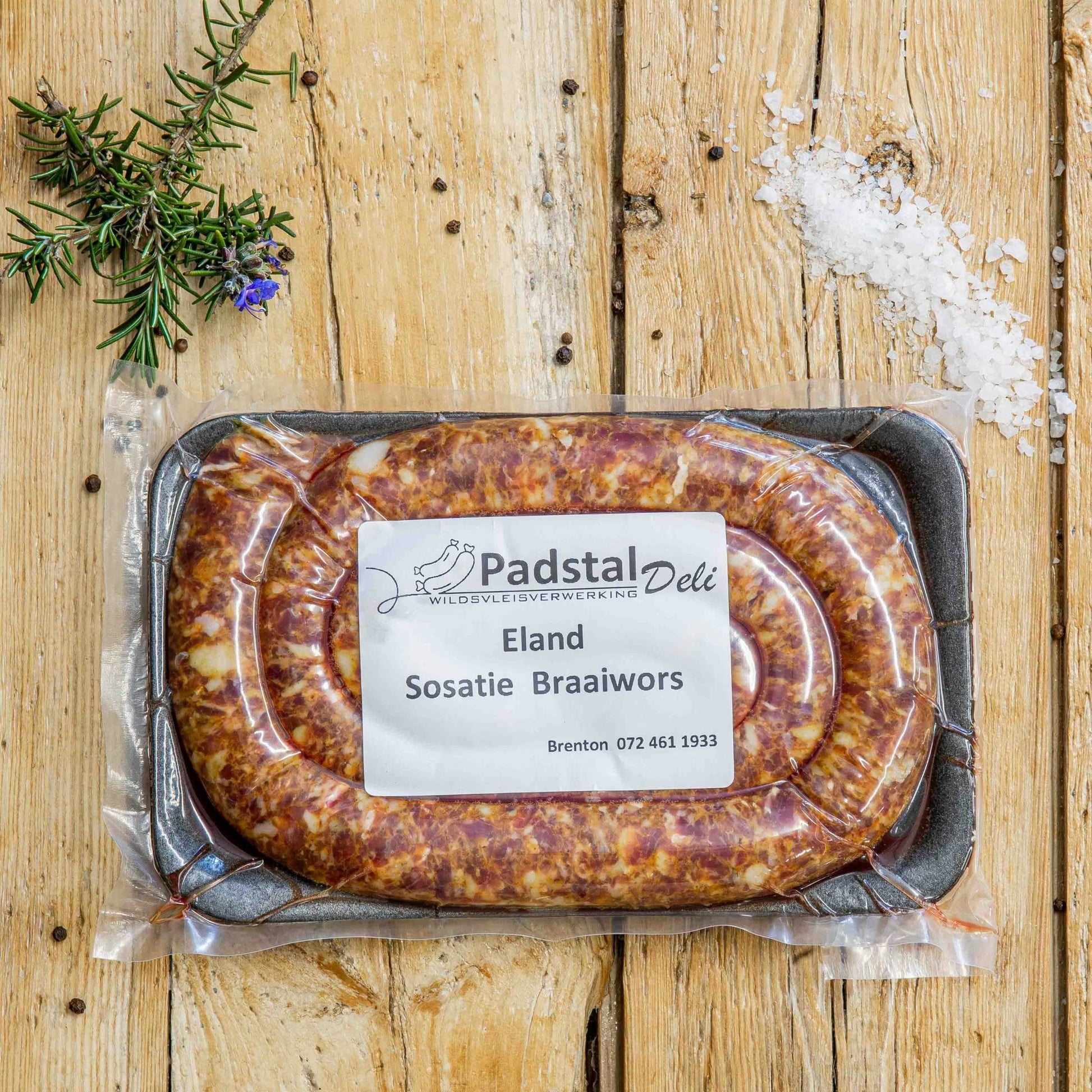 Padstal Deli vacuum-sealed Eland Sosatie Braaiwors packaged on a black tray, styled on a light wooden background with coarse sea salt, black peppercorns, and fresh rosemary.