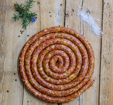 Padstal Deli Eland Sosatie Braaiwors coiled in a tight spiral on a light rustic wooden background, decorated with coarse sea salt, black peppercorns, and fresh rosemary.