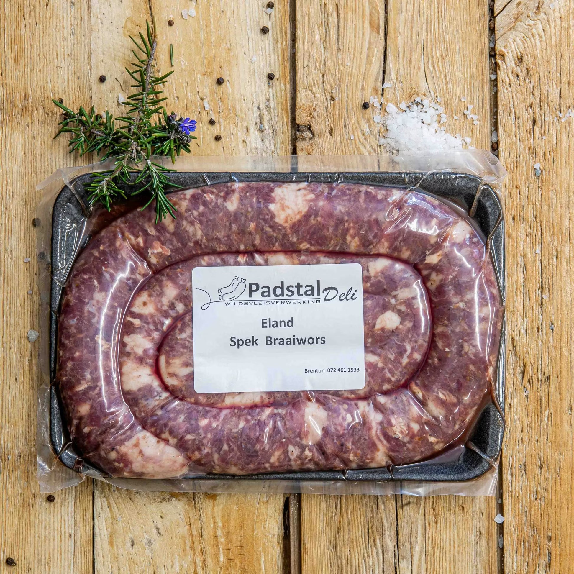 Padstal Deli vacuum-sealed Eland Spek Braaiwors packaged on a black tray, styled on a light wooden background with coarse sea salt, black peppercorns, and fresh rosemary.