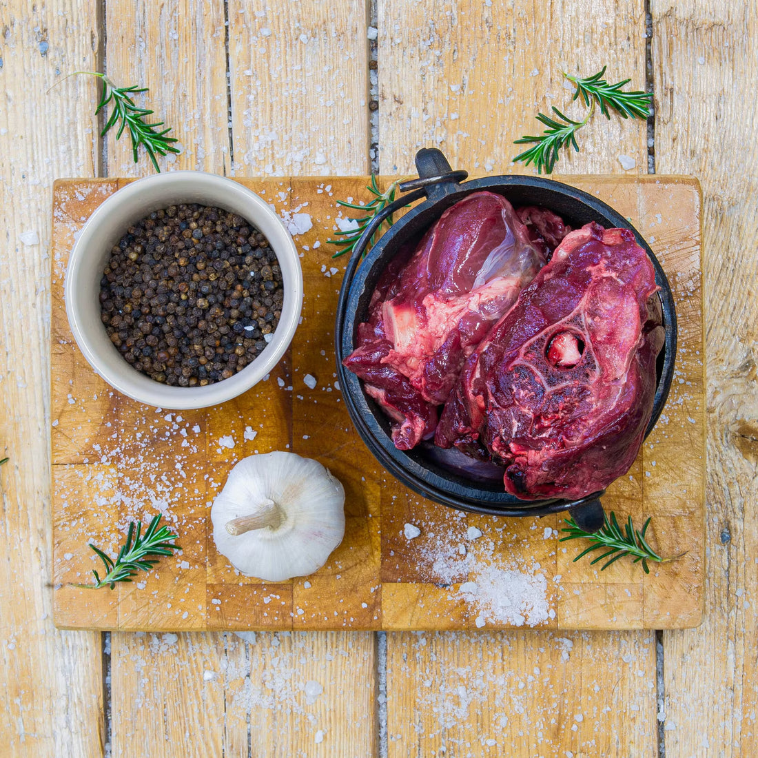 Premium raw game potjie meat pieces packed inside a small black cast iron potjie pot on a wooden cutting board, garnished with a whole garlic bulb, black peppercorns, and fresh rosemary.