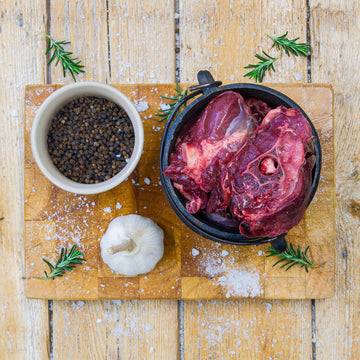 Premium raw game potjie meat pieces packed inside a small black cast iron potjie pot on a wooden cutting board, garnished with a whole garlic bulb, black peppercorns, and fresh rosemary.