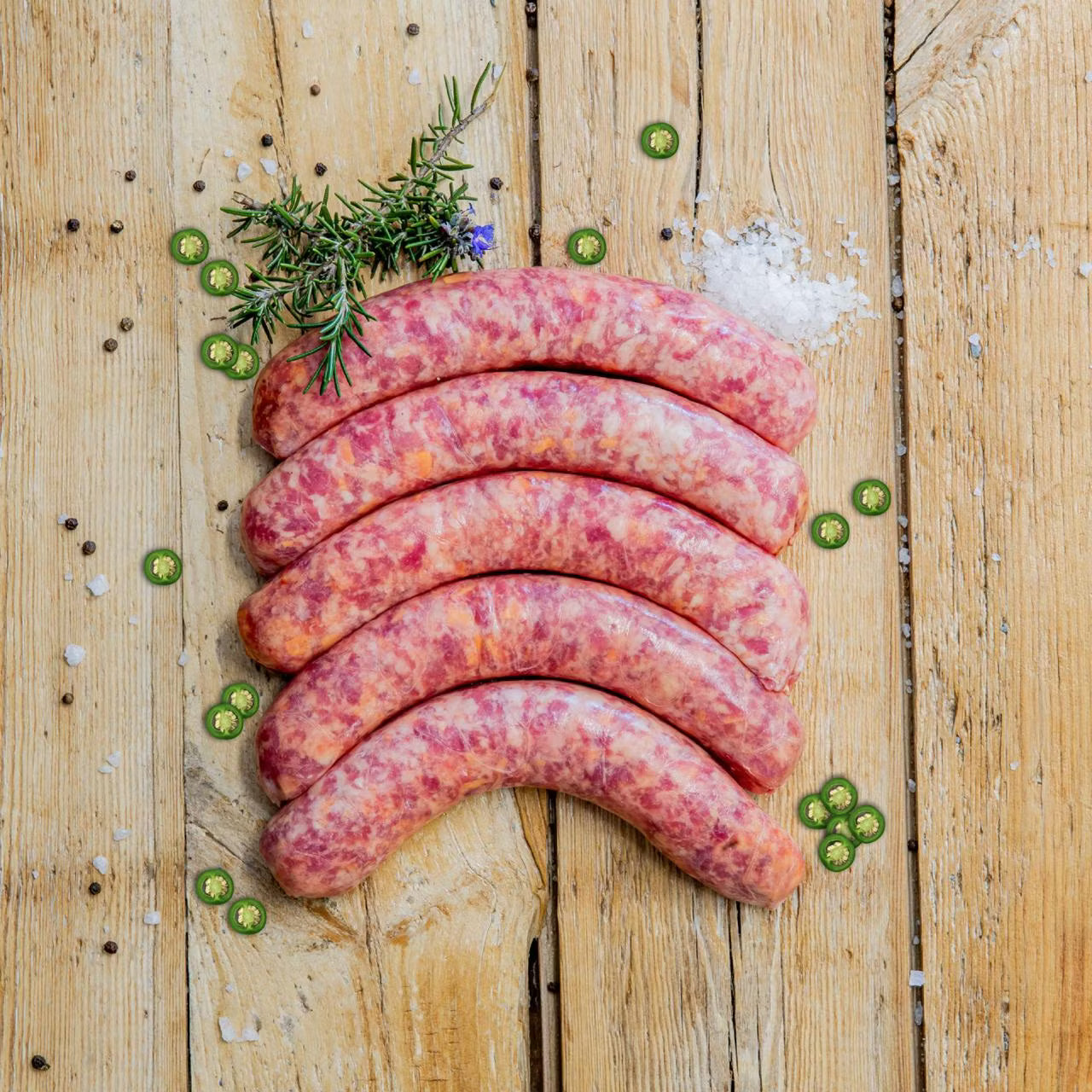 Padstal Deli premium raw Jalapeño Kaaswors (cheese sausages) arranged on a light rustic wooden background, decorated with fresh sliced jalapeño rings, coarse sea salt, and a sprig of rosemary.