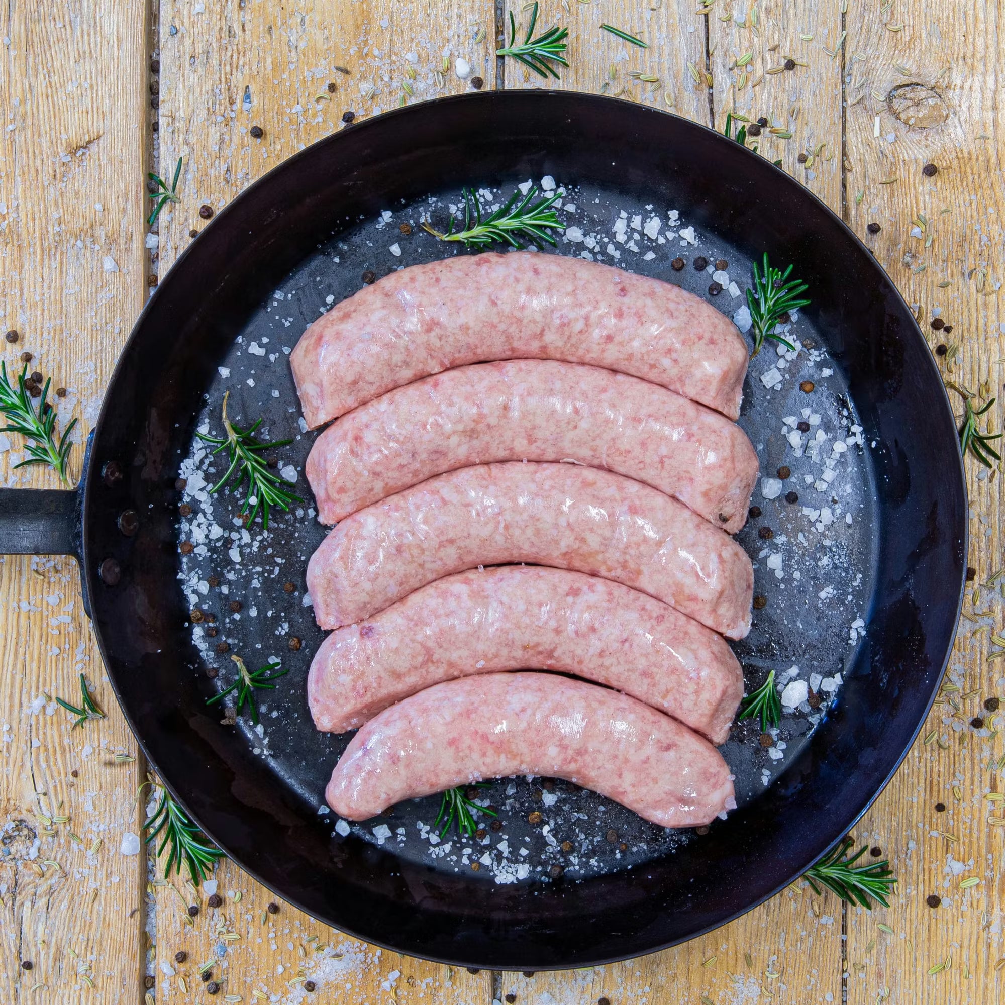 Premium Padstal Deli raw Pork Bangers arranged in a black cast-iron skillet, garnished with coarse sea salt, black peppercorns, and fresh rosemary sprigs.
