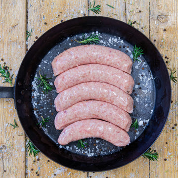Premium Padstal Deli raw Pork Bangers arranged in a black cast-iron skillet, garnished with coarse sea salt, black peppercorns, and fresh rosemary sprigs.