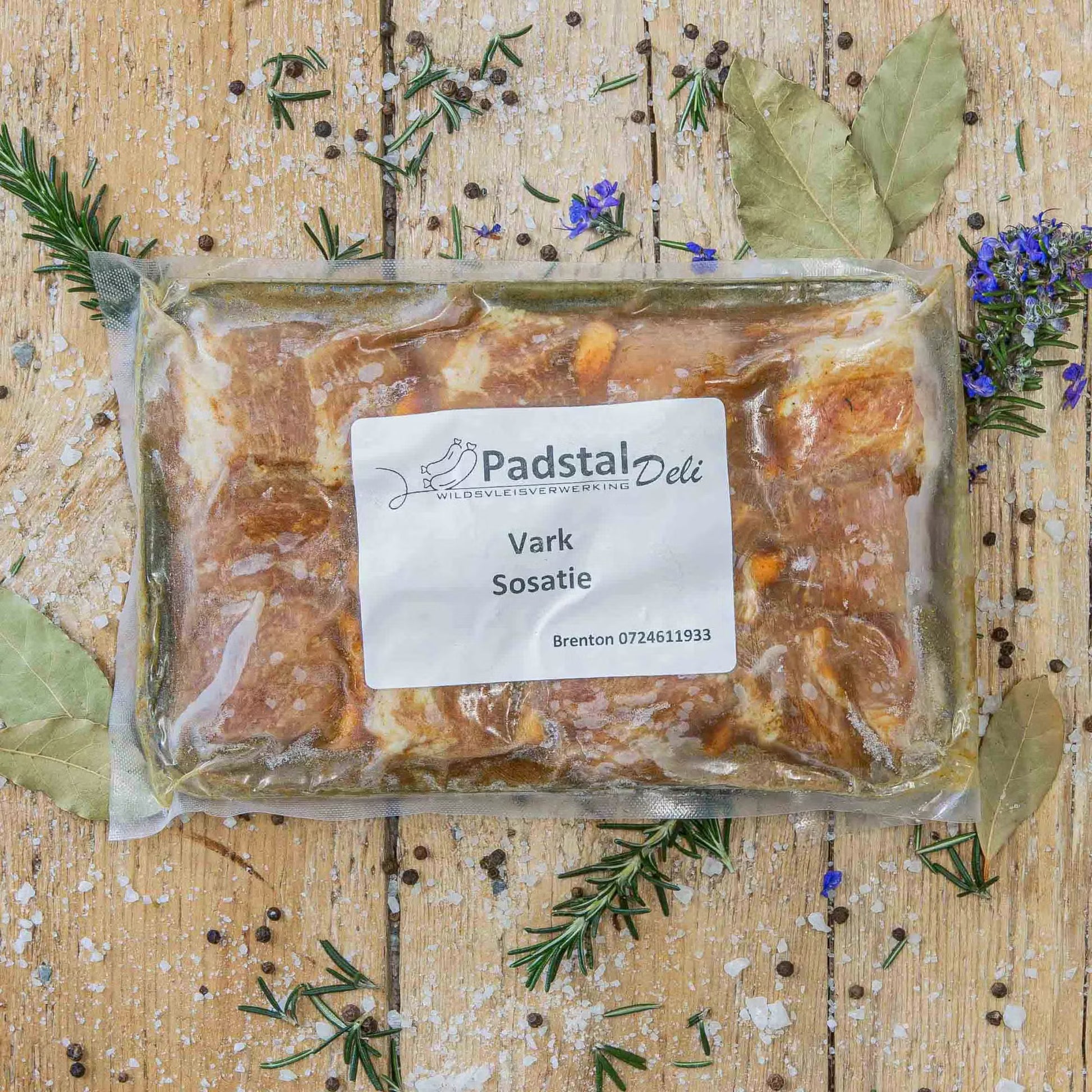 Padstal Deli vacuum-sealed premium Vark Sosatie pack resting on a rustic wooden background, decorated with fresh bay leaves, rosemary, coarse salt, and peppercorns.