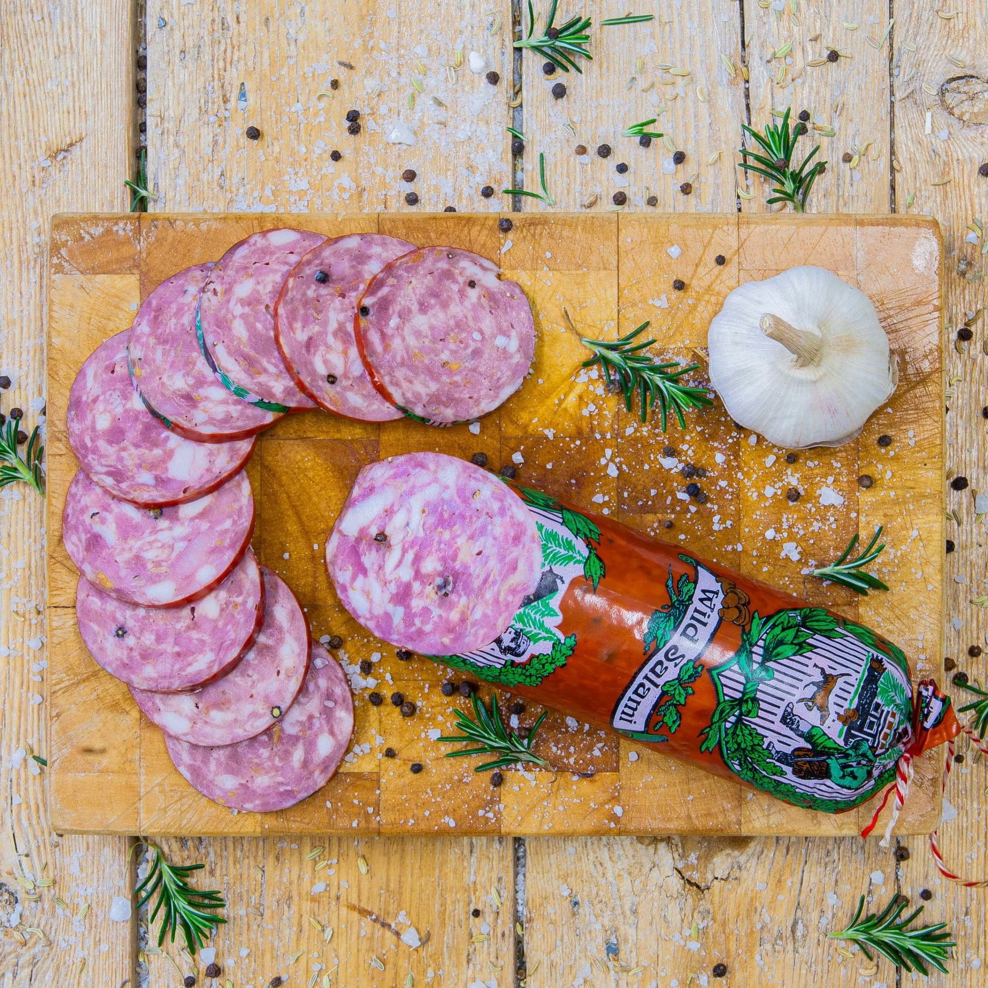 Padstal Deli premium Wild Salami stick with gourmet slices arranged on a wooden cutting board, garnished with a whole garlic bulb, coarse salt, and fresh rosemary.