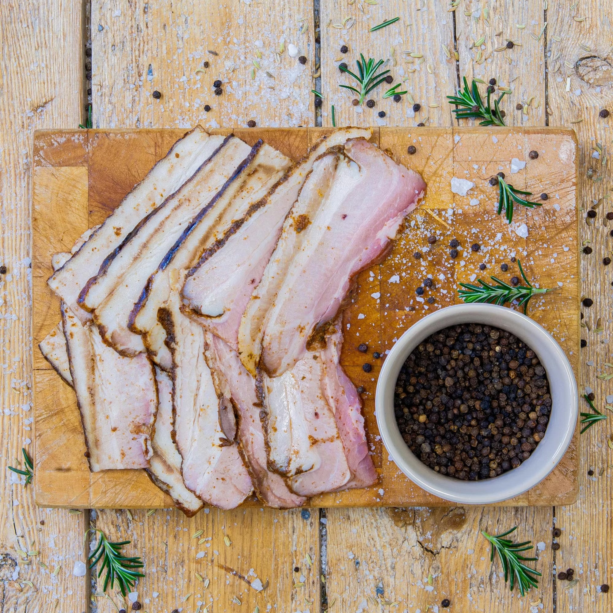 Padstal Deli premium Smoked Rashers sliced and arranged on a wooden cutting board next to a small bowl of whole black peppercorns, coarse salt, and fresh rosemary.