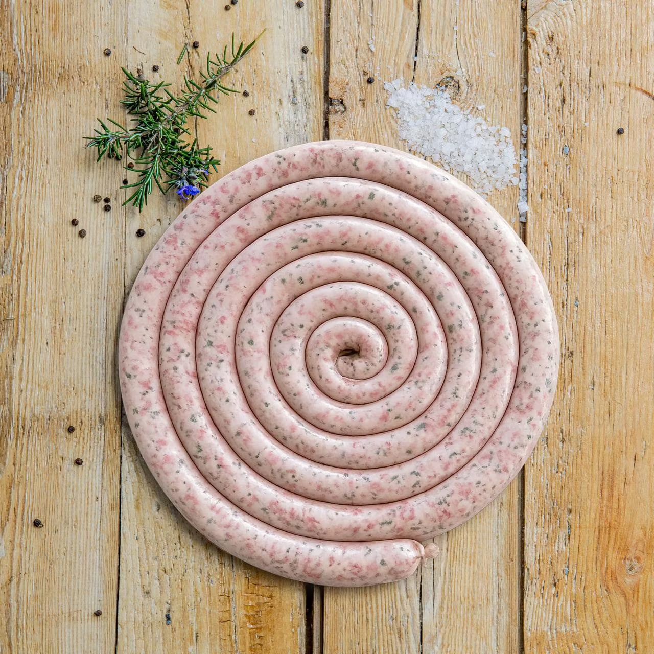 Traditional Padstal Deli pork braaiwors coiled in a perfect spiral on a wooden surface, garnished with coarse salt, peppercorns, and fresh rosemary.