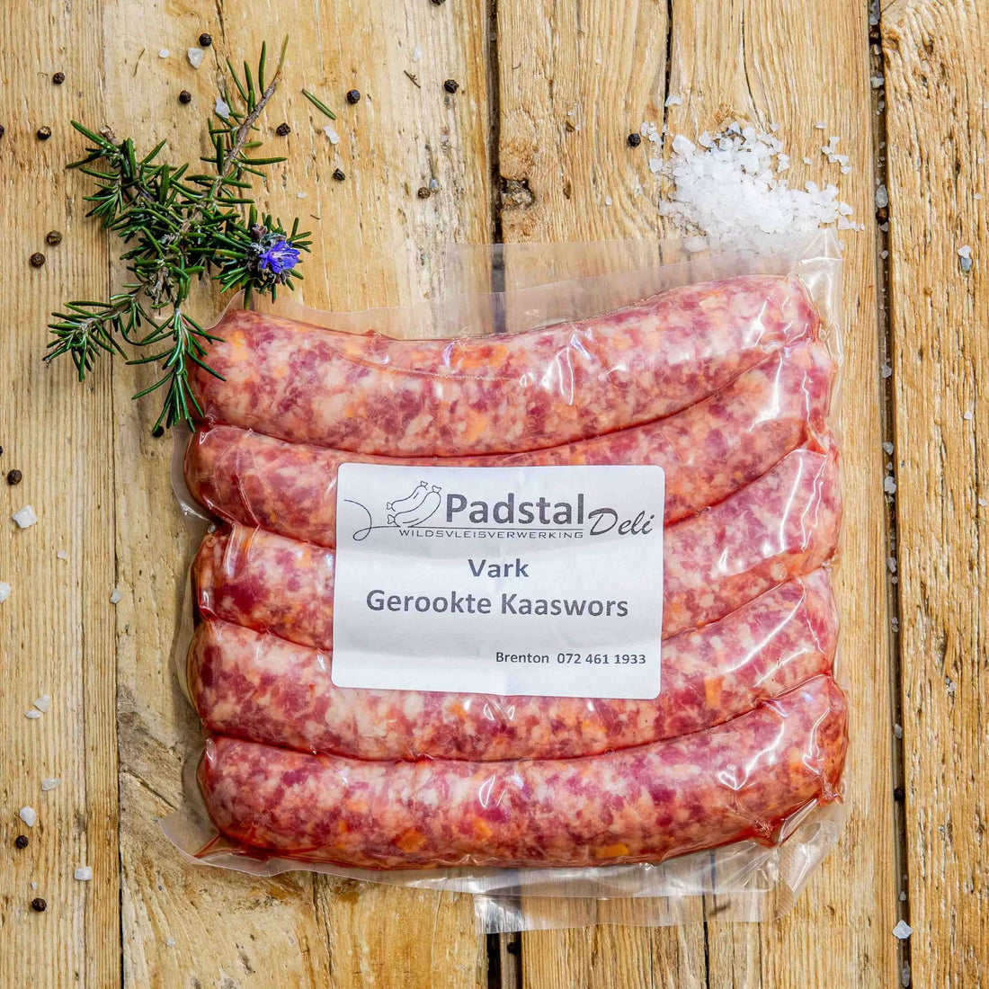Padstal Deli vacuum-sealed premium Vark Gerookte Kaaswors (smoked pork cheese sausage) pack resting on a rustic wooden background with coarse salt, peppercorns, and fresh rosemary.