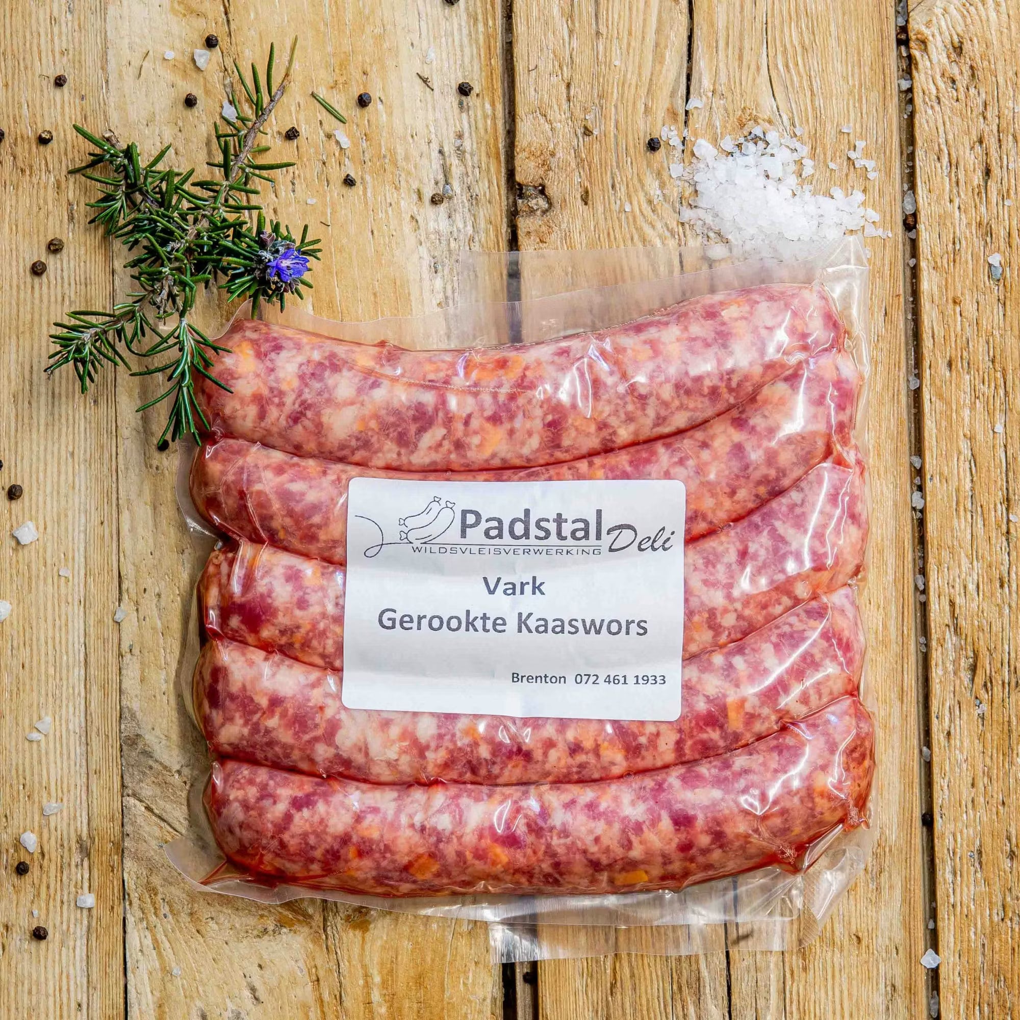 Padstal Deli vacuum-sealed premium Vark Gerookte Kaaswors (smoked pork cheese sausage) pack resting on a rustic wooden background with coarse salt, peppercorns, and fresh rosemary.
